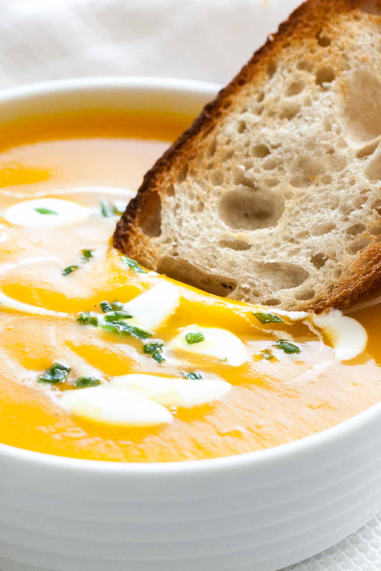Close-up of a toasted slice of bread being dipped into butternut squash soup with dollops of sour cream and chives in a white bowl.