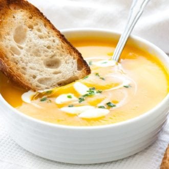 A white bowl with butternut squash soup, with dollops of sour cream and chives, a spoon in it and a toasted slice of bread. It is sitting on a white dish towel next to some toasted slices of bread.
