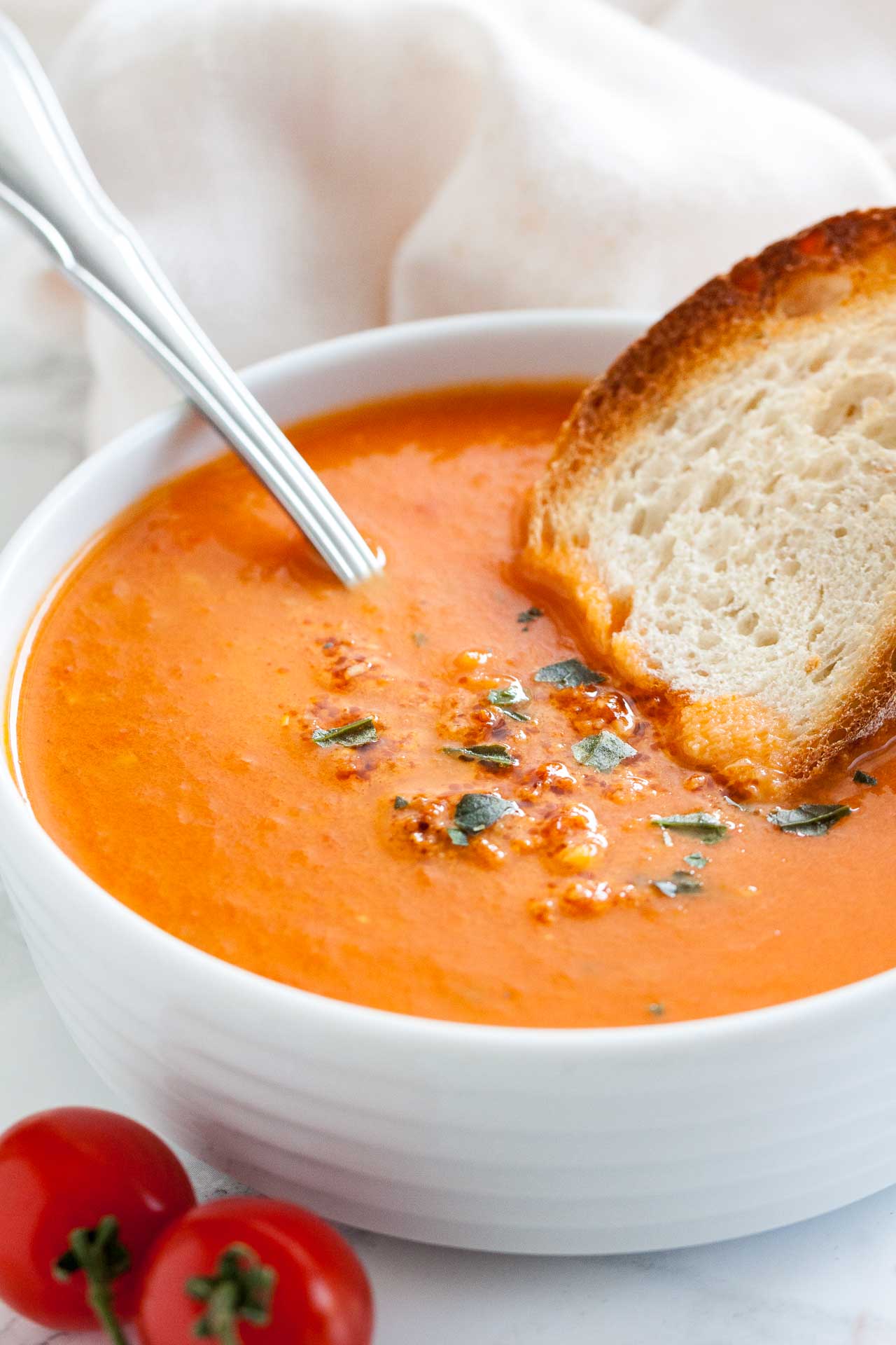 A white bowl of roasted tomato soup garnished with basil, with a spoon and a toasted slice of bread on a white dishtowel.