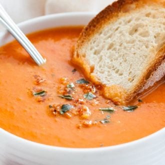 A white bowl of roasted tomato soup garnished with basil, with a spoon and a toasted slice of bread on a white dishtowel.