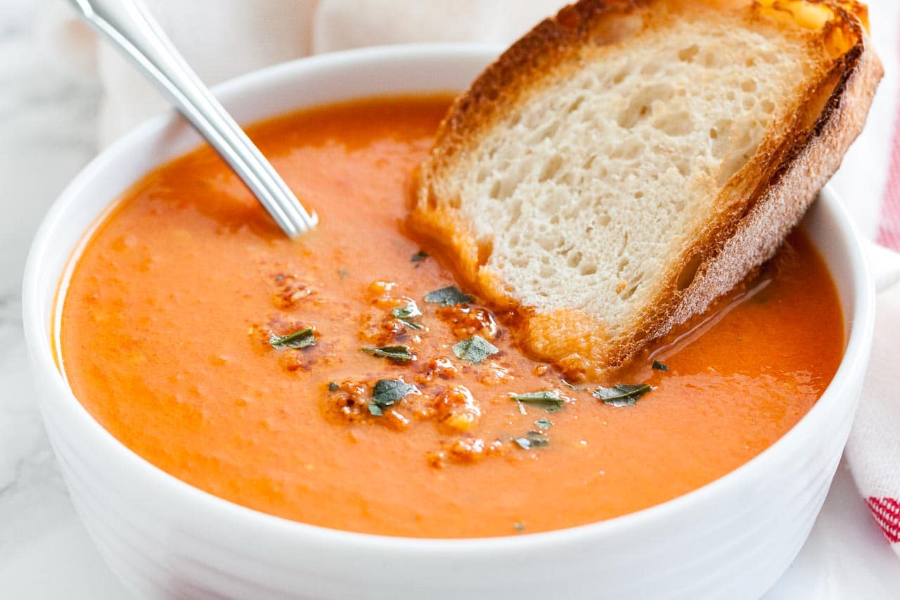 A white bowl of roasted tomato soup garnished with basil, with a spoon and a toasted slice of bread on a white dishtowel.