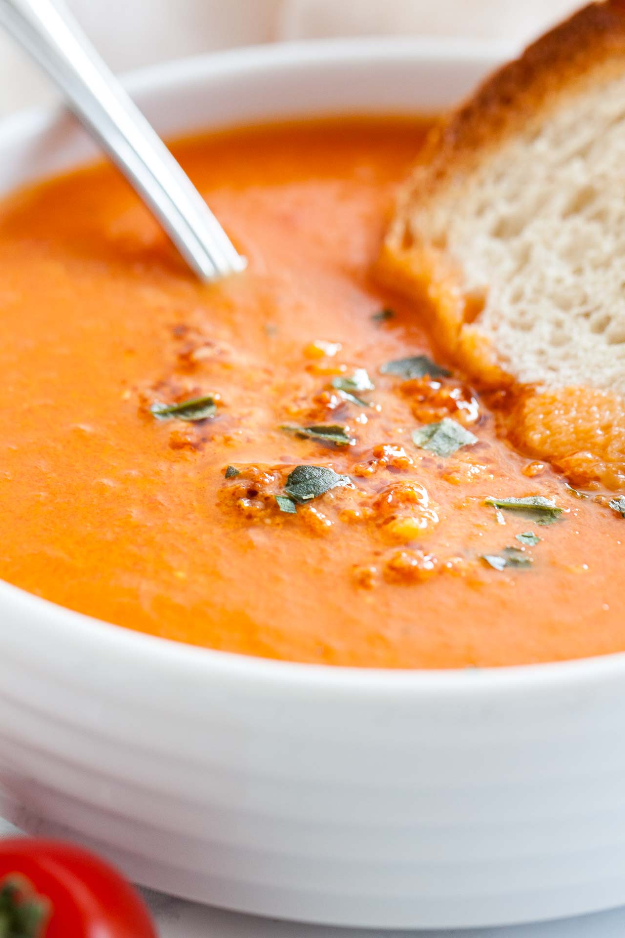 Close-up of a white bowl of roasted tomato soup garnished with basil, with a spoon and a toasted slice of bread.