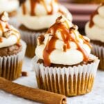 Pumpkin cupcakes with brown butter frosting, topped with caramel sauce and walnut pieces on a grey tablecloth with a cinnamon stick.