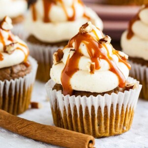 Pumpkin cupcakes with brown butter frosting, topped with caramel sauce and walnut pieces on a grey tablecloth with a cinnamon stick.