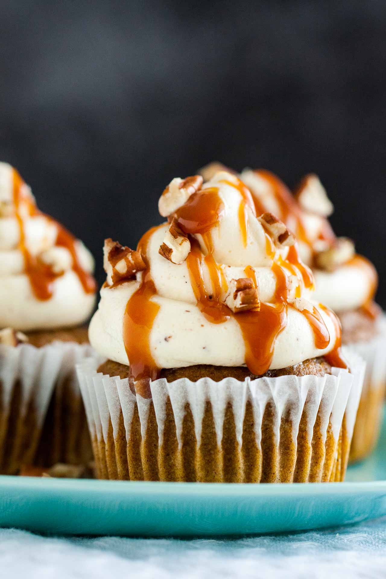 Pumpkin cupcakes with brown butter frosting, topped with caramel sauce and walnut pieces on a teal plate.