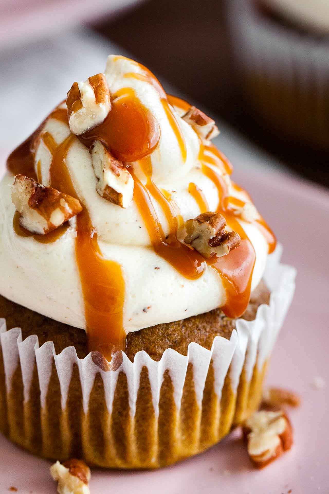 Close-up of a pumpkin cupcake with brown butter frosting, topped with caramel sauce and walnut pieces on a pink plate.