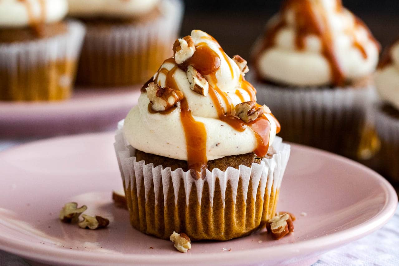 A pumpkin cupcake with brown butter frosting, topped with caramel sauce and walnut pieces on a pink plate garnished with walnut pieces. There are more cupcakes in the background.