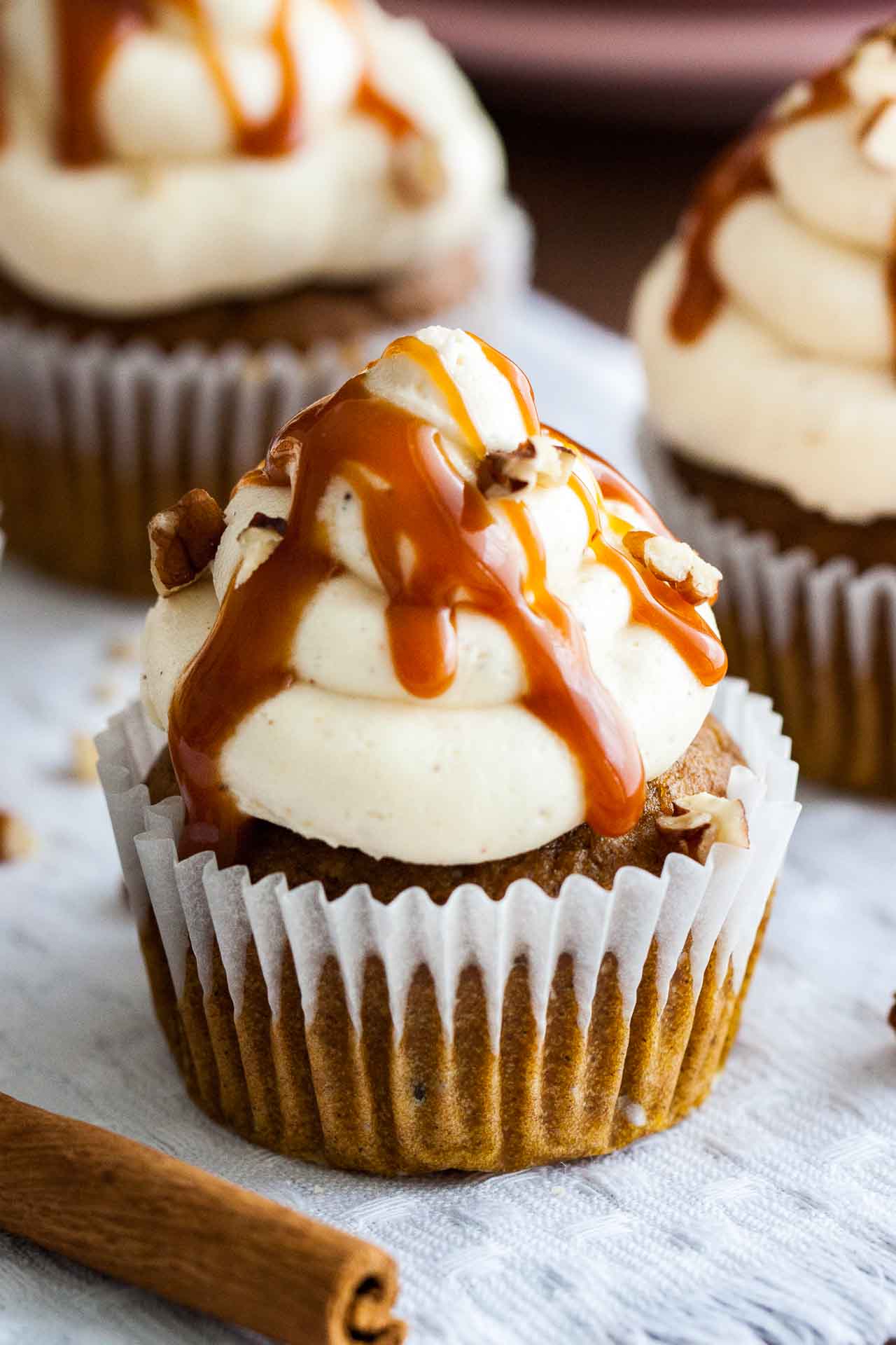 Close-up of a pumpkin cupcake with brown butter frosting, topped with caramel sauce and walnut pieces on a grey tablecloth with a cinnamon stick.