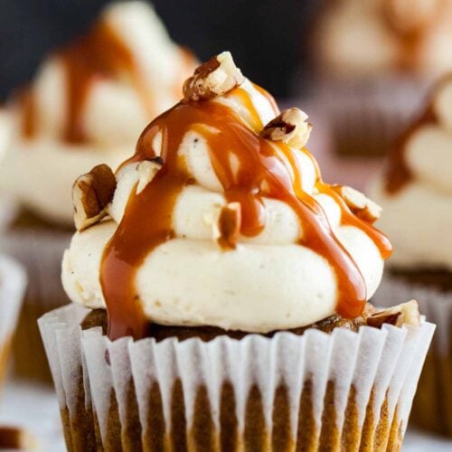 Close-up of a pumpkin cupcake with brown butter frosting, topped with caramel sauce and walnut pieces on a grey tablecloth with a cinnamon stick.