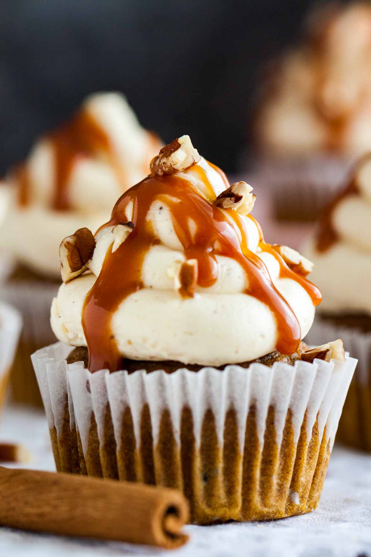 Close-up of a pumpkin cupcake with brown butter frosting, topped with caramel sauce and walnut pieces on a grey tablecloth with a cinnamon stick.