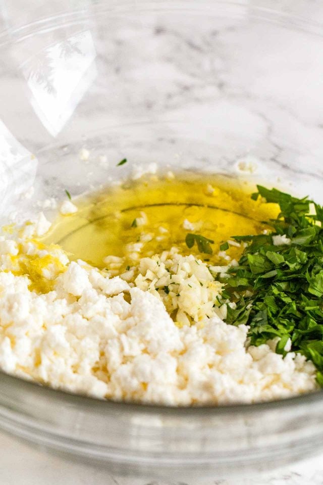 A glass bowl with olive oil, minced garlic, lemon juice, feta and chopped parsley on a marble surface.