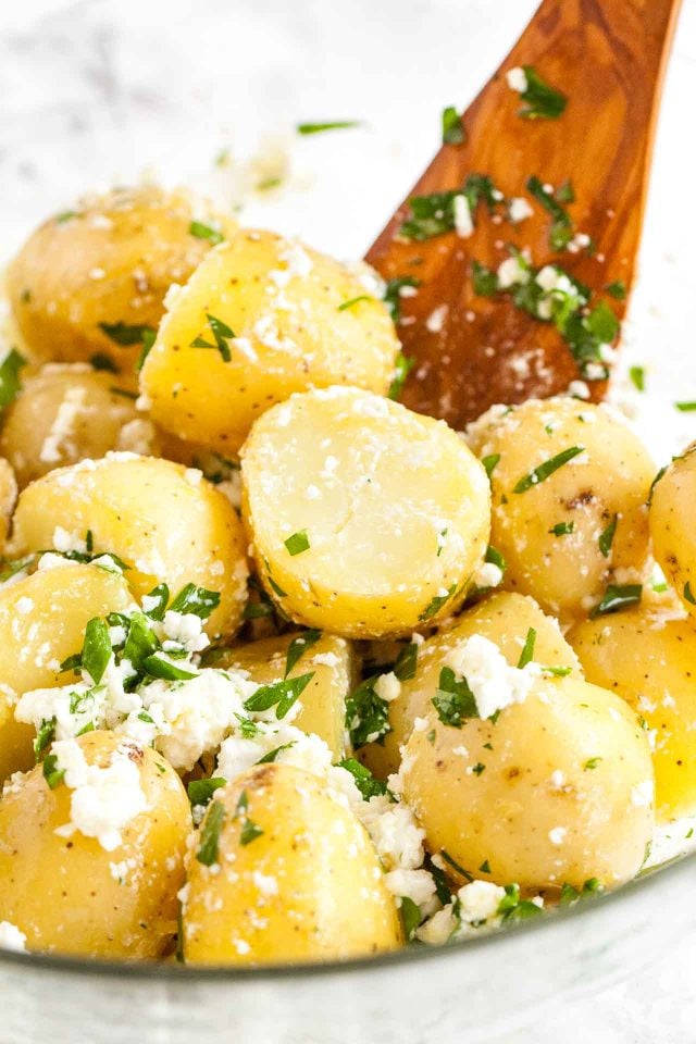 Close-up of a glass bowl of potatoes with feta and parsley with a wooden spatula.