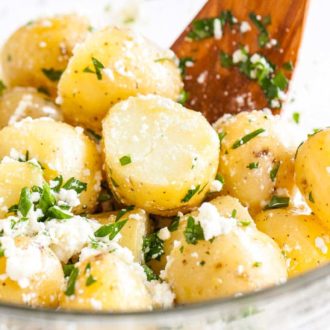 A glass bowl of potatoes with feta and parsley with a wooden spatula.
