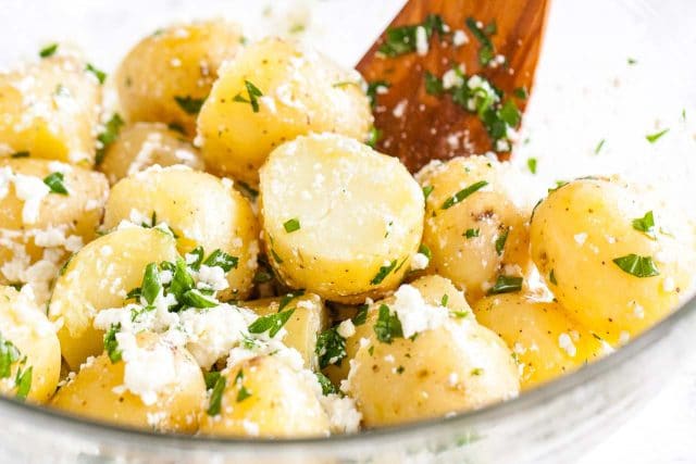 A glass bowl of potatoes with feta and parsley with a wooden spatula.