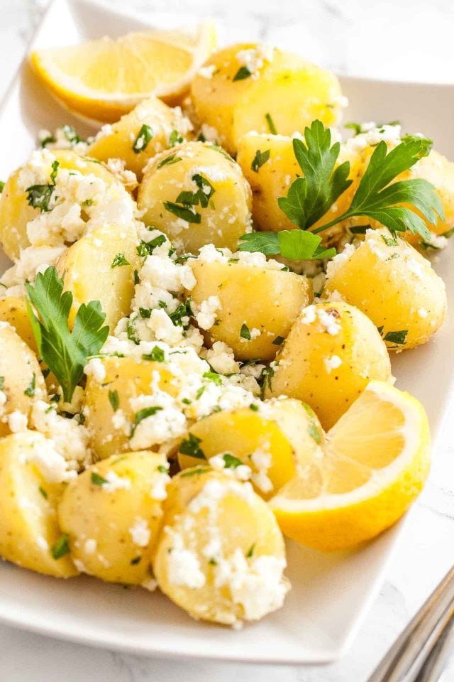 Close-up of a rectangular plate with potatoes with feta and parsley with a wedge of lemon on a marble surface.