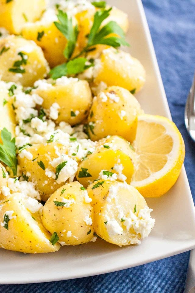 A rectangular plate with potatoes with feta and parsley with a wedge of lemon on a blue surface with a spoon next to it.