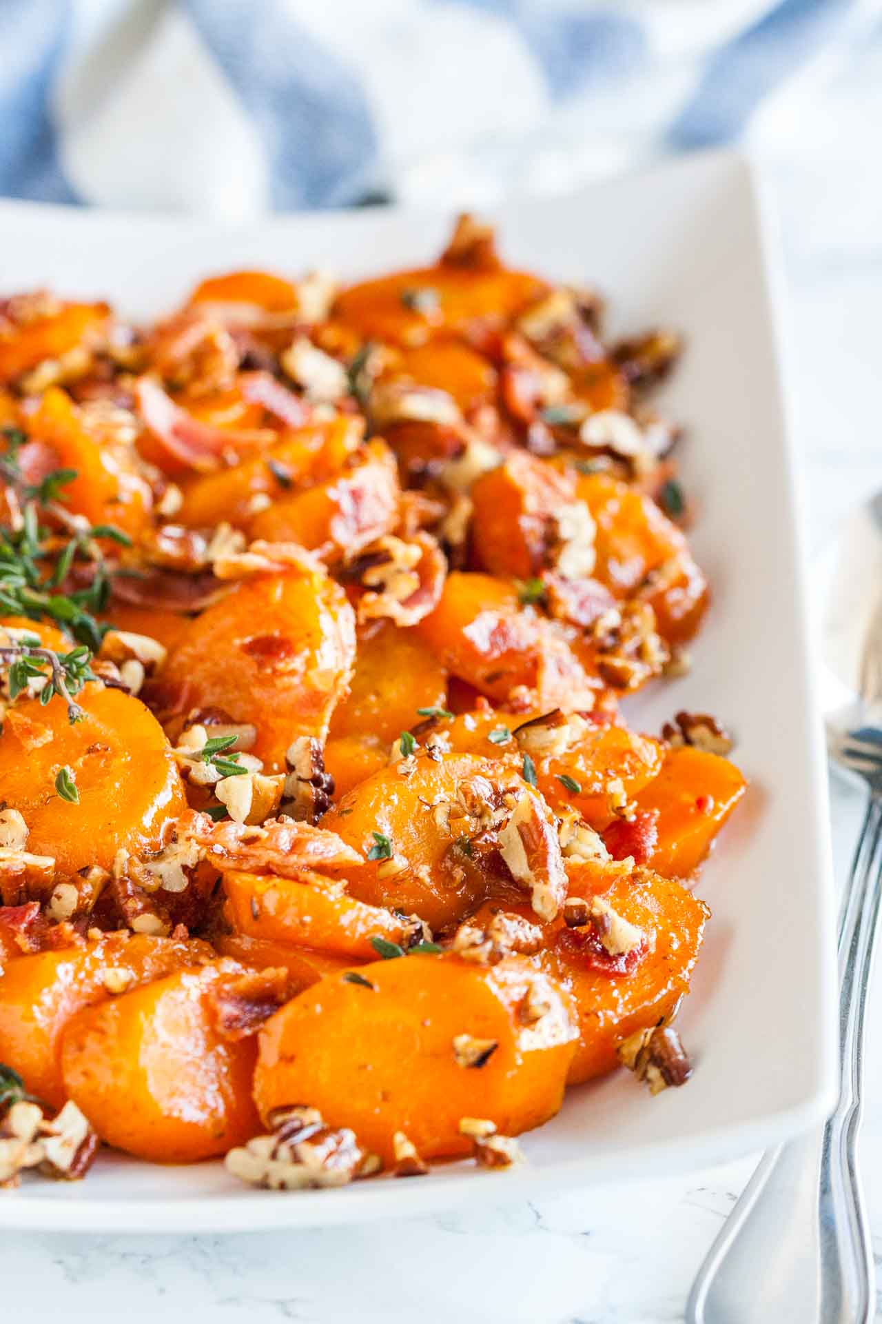 A rectangular white plate of brown sugar-glazed carrots with bacon and pecans garnished with thyme on a marble surface with a spoon next to it.