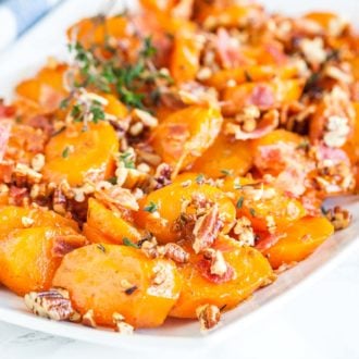 A rectangular white plate of brown sugar-glazed carrots with bacon and pecans garnished with thyme.