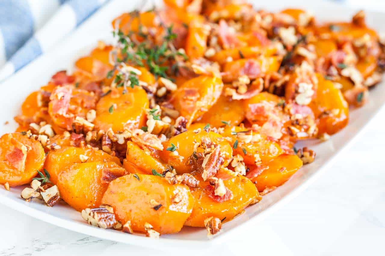 A rectangular white plate of brown sugar-glazed carrots with bacon and pecans garnished with thyme.