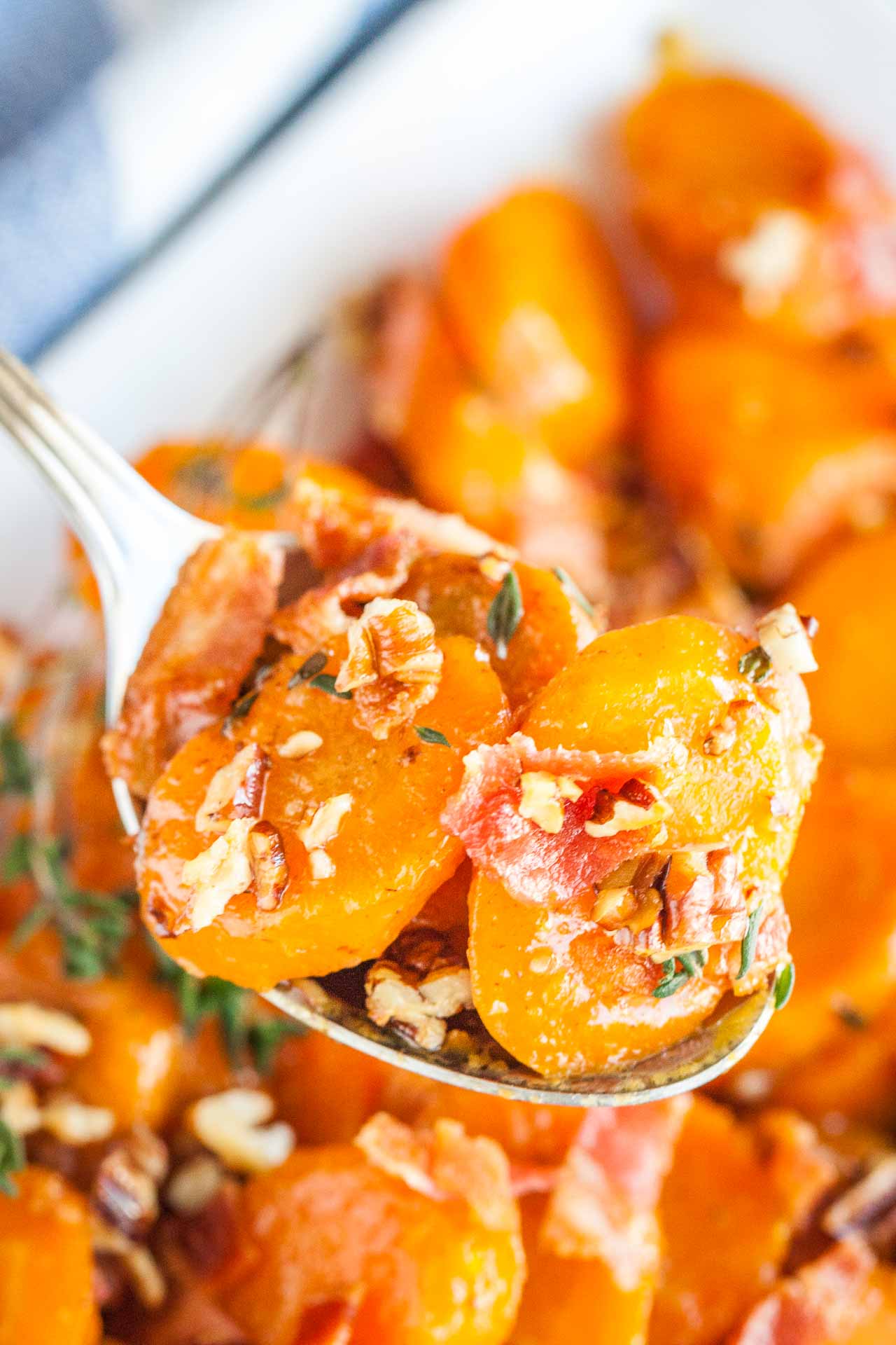 A spoon holding up some brown sugar-glazed carrots with bacon and pecans above a white bowl of carrots.