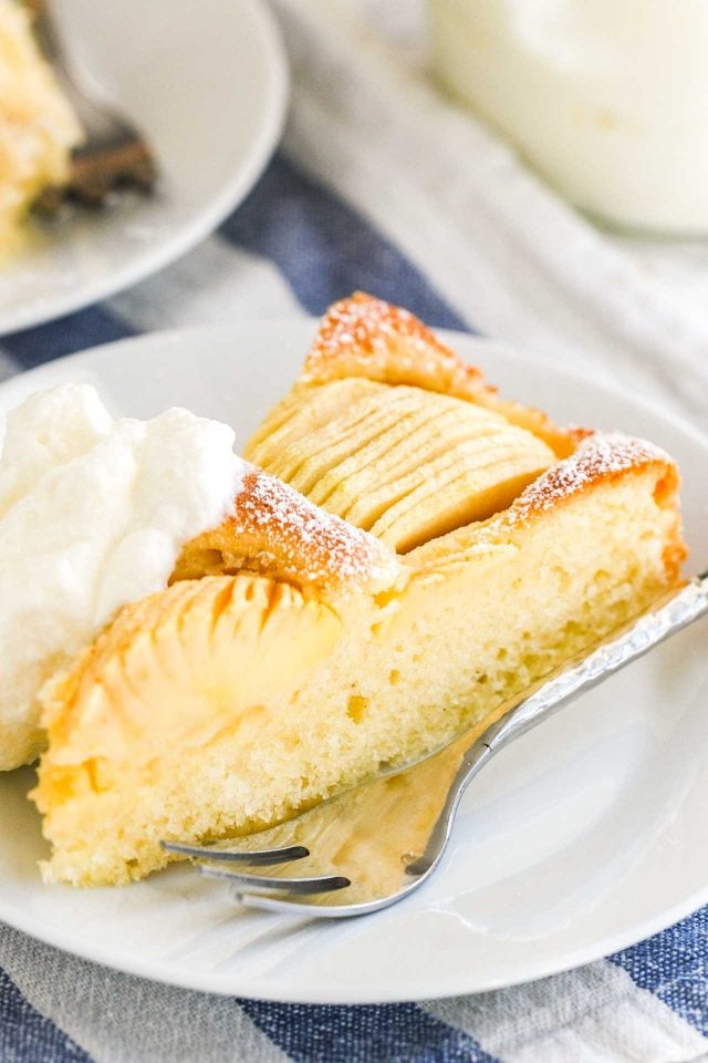 A slice of cake with apple quarters sunken into the top on a white plate, with whipped cream and a fork.