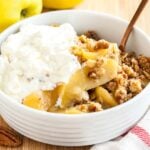 A white bowl of apple crisp, topped with whipped cream and a bronze spoon in it on a bamboo cutting board with apples and a white and red dishtowel.