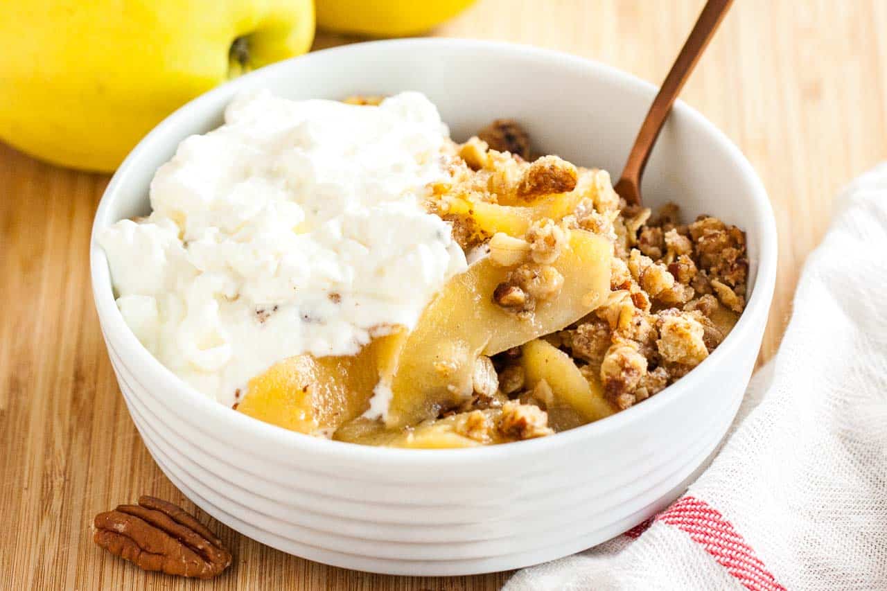 A white bowl of apple crisp, topped with whipped cream and a bronze spoon in it on a bamboo cutting board with apples and a white and red dishtowel.