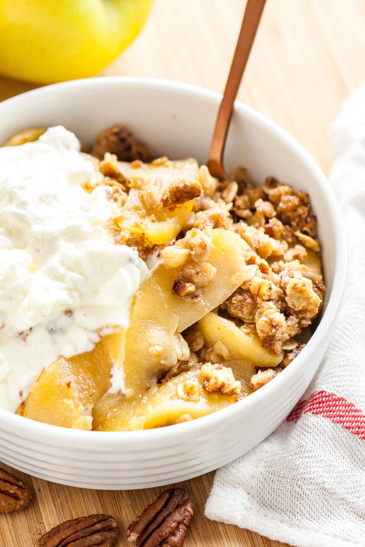 A white bowl of apple crisp, topped with whipped cream and a bronze spoon in it on a bamboo cutting board with apples and a white and red dishtowel.