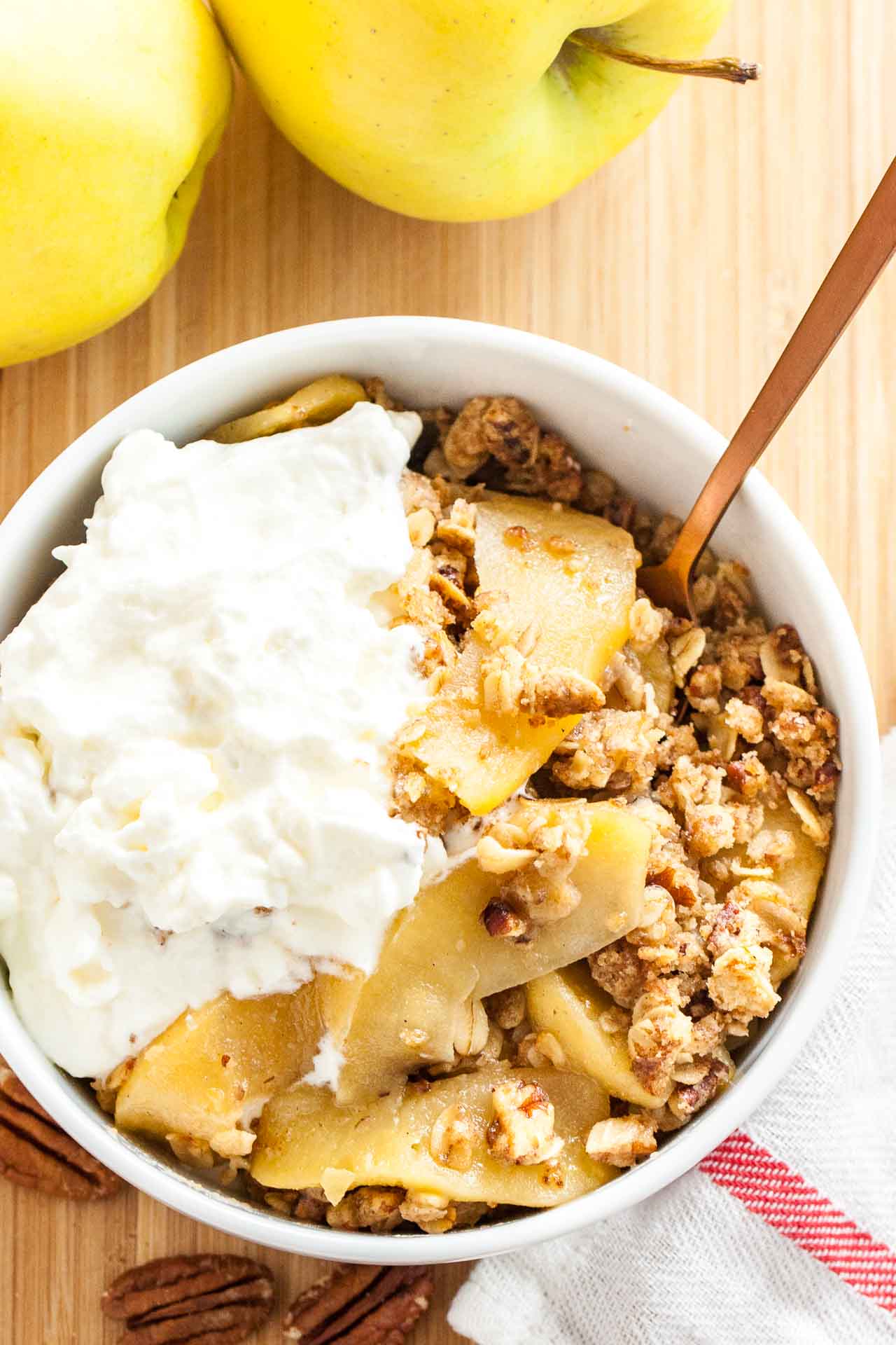 Top-down shot of a white bowl of apple crisp, topped with whipped cream and a bronze spoon in it on a bamboo cutting board with apples and a white and red dishtowel.