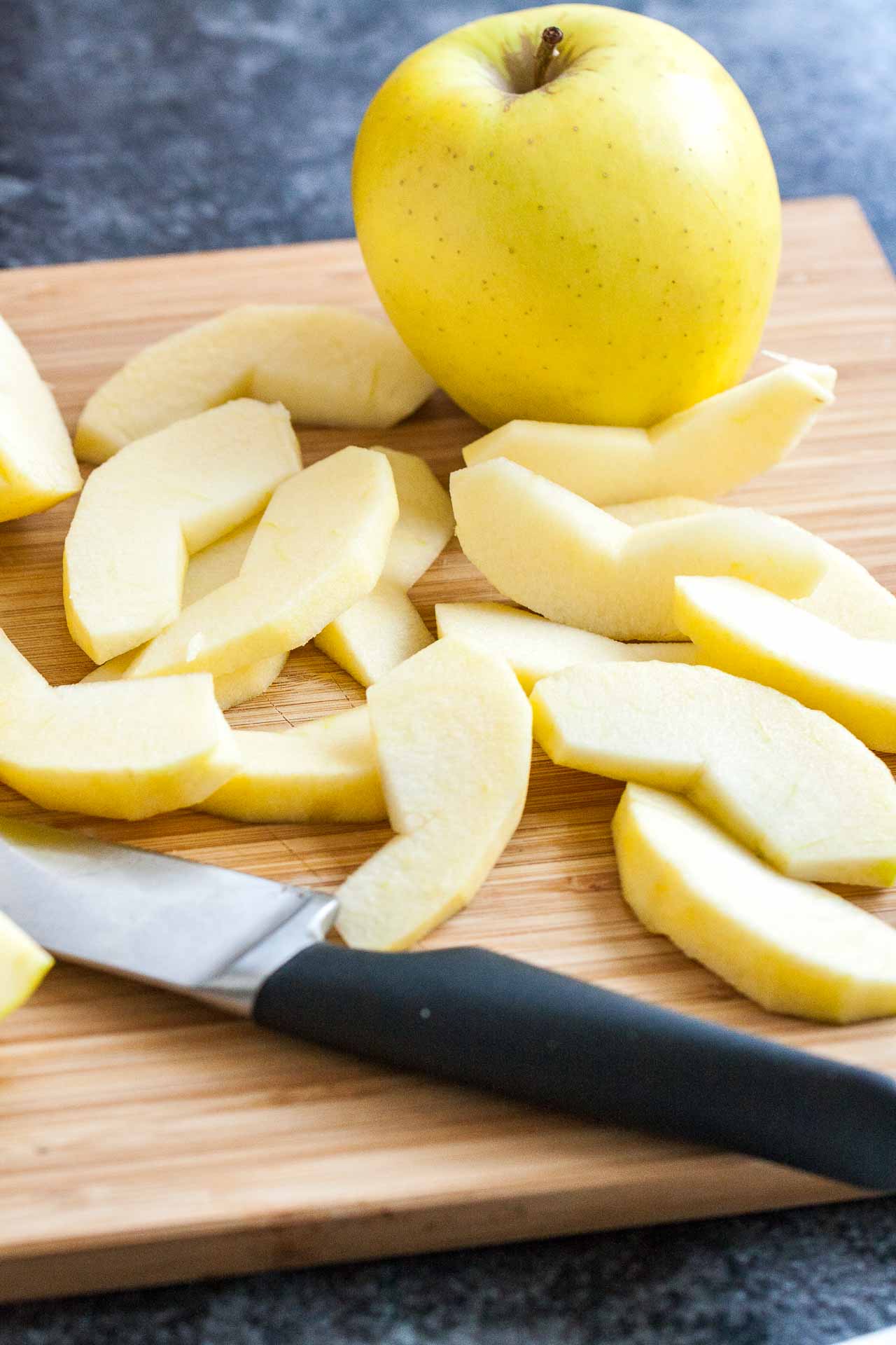 A bamboo cutting board with peeled apple wedges, a whole apple and a knife.