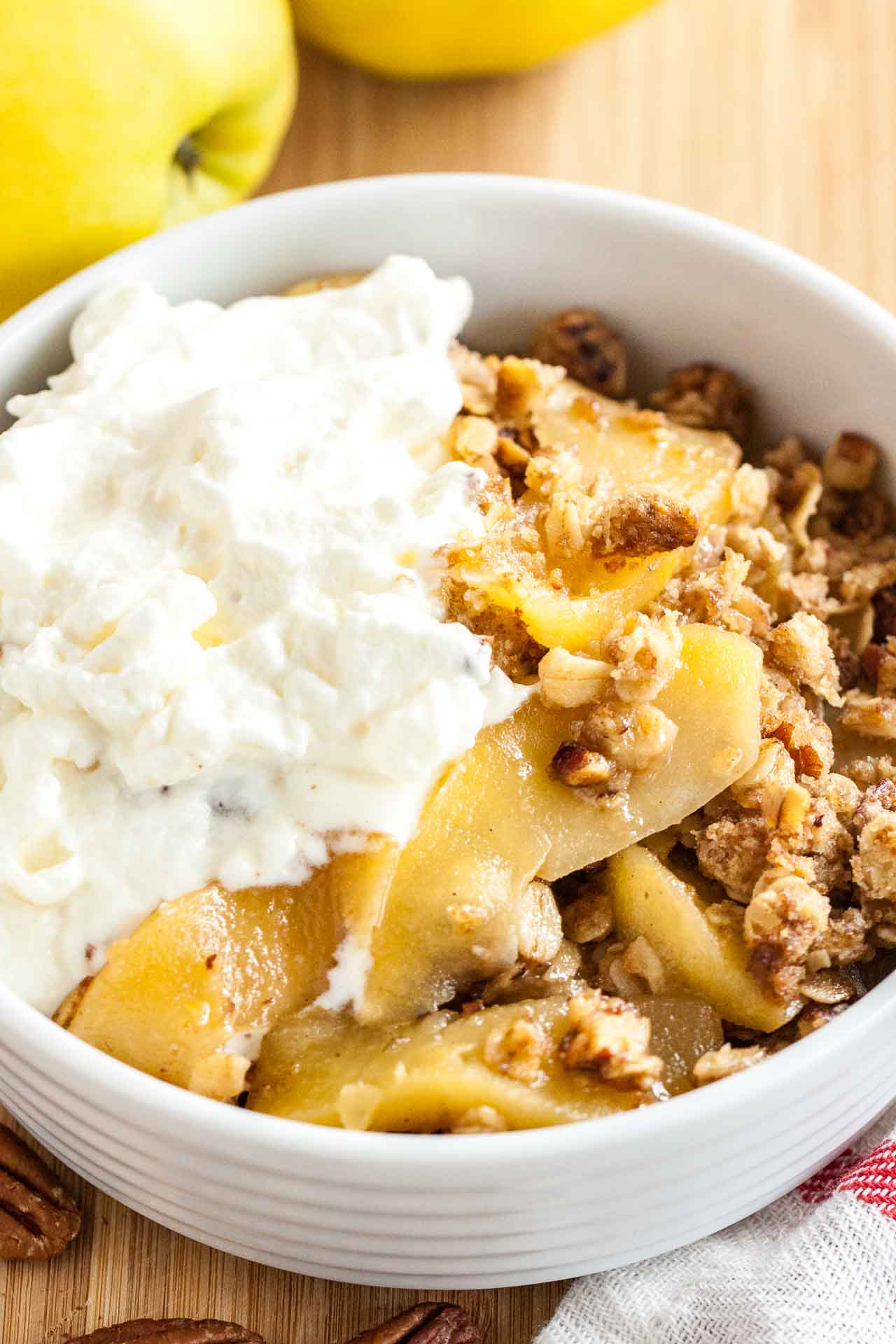 A white bowl of apple crisp, topped with whipped cream it on a bamboo cutting board with apples and a white and red dishtowel.