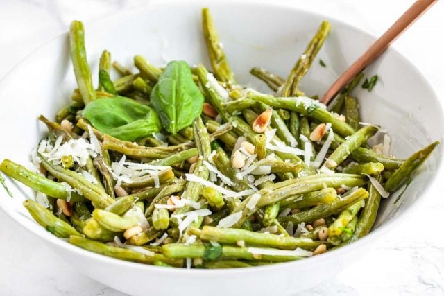 A white bowl of oven-roasted green beans topped with roasted pine nuts and Parmigiano, garnished with a basil leaves.