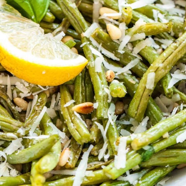 Close up of oven-roasted green beans topped with roasted pine nuts and Parmigiano, garnished with a basil leaf and a wedge of lemon.