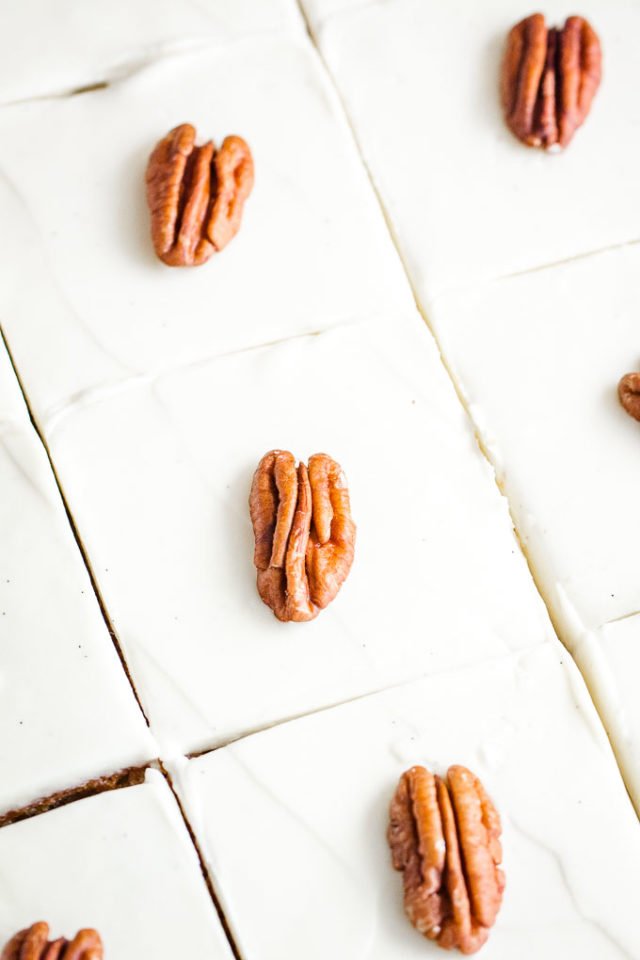 Top-down close-up shot of a sheet cake topped with white frosting and pecan nuts, cut into squares.