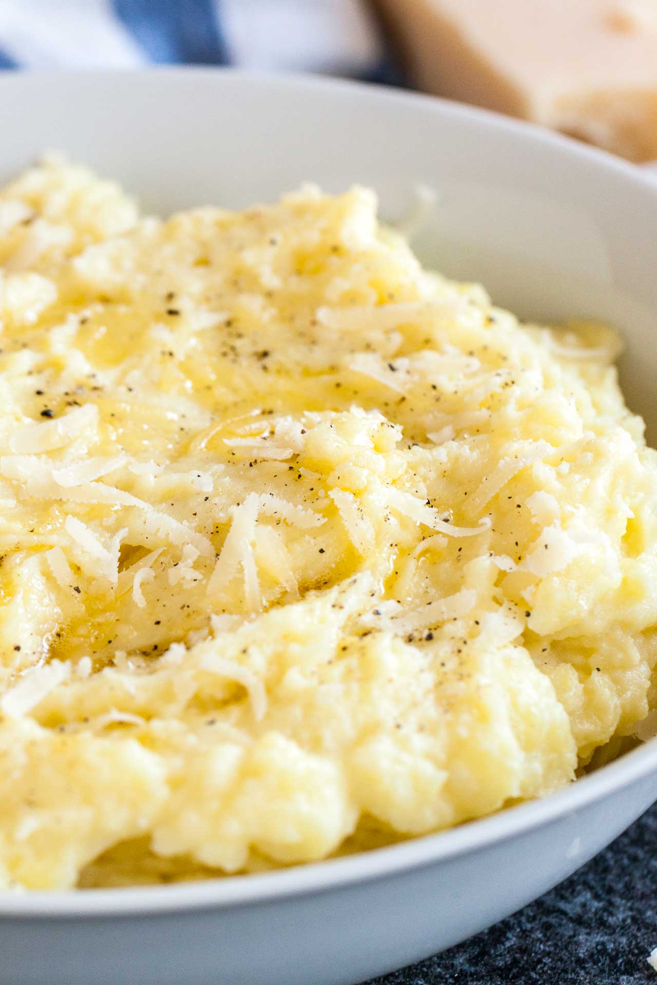 A white bowl of roasted garlic mashed potatoes topped with Parmigiano cheese and pepper. There\'s a white and blue dishtowel and a block of Parmigiano in the background.