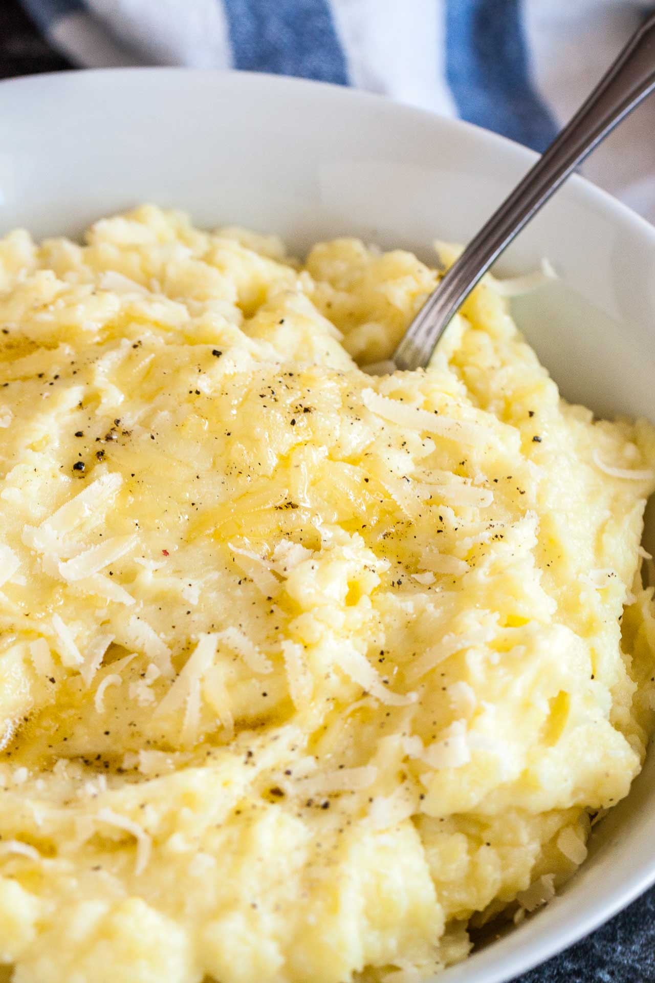 Close-up of a white bowl of roasted garlic mashed potatoes topped with Parmigiano cheese and pepper with a spoon in it. There\'s a white and blue dishtowel in the background.