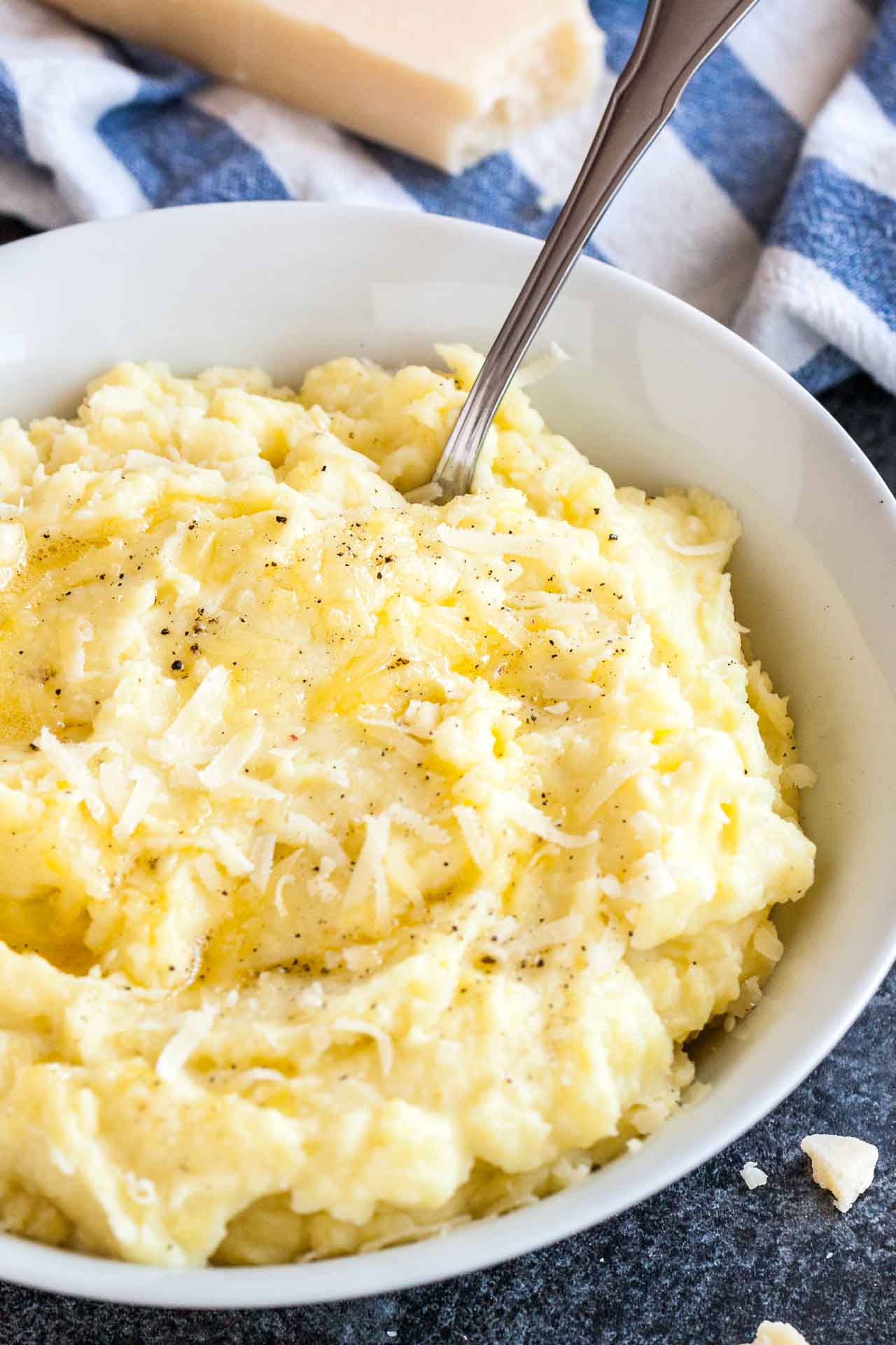 A white bowl of roasted garlic mashed potatoes topped with Parmigiano cheese and pepper with a spoon in it. There\'s a white and blue dishtowel and a block of Parmigiano in the background.