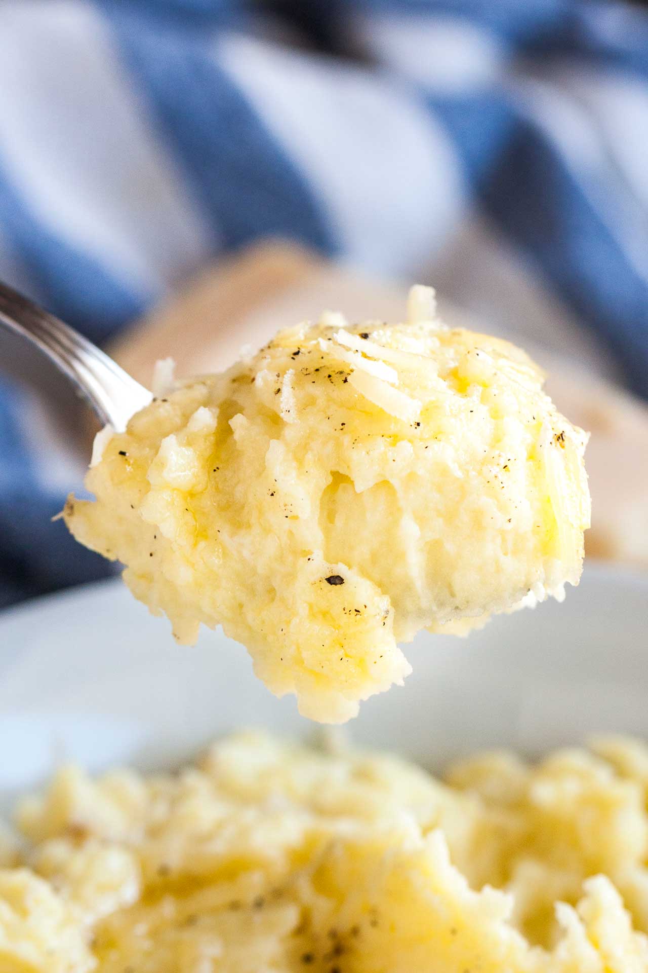 A spoon holding up a blob of roasted garlic mashed potatoes above a white bowl of mashed potatoes with a white and blue dish towel and a block of Parmigiano in the background.