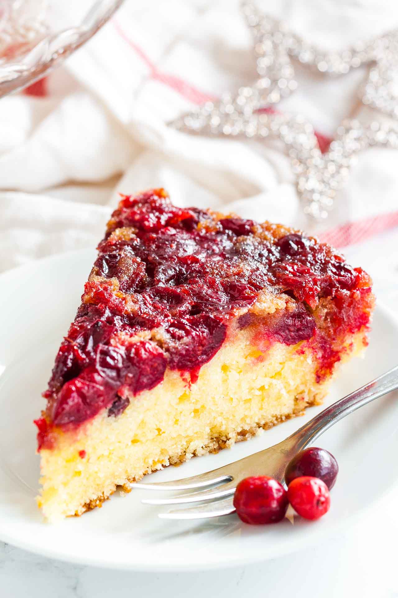 A slice of upside-down cranberry cake on a white plate with a fork, garnished with cranberries and Christmas stars in the background