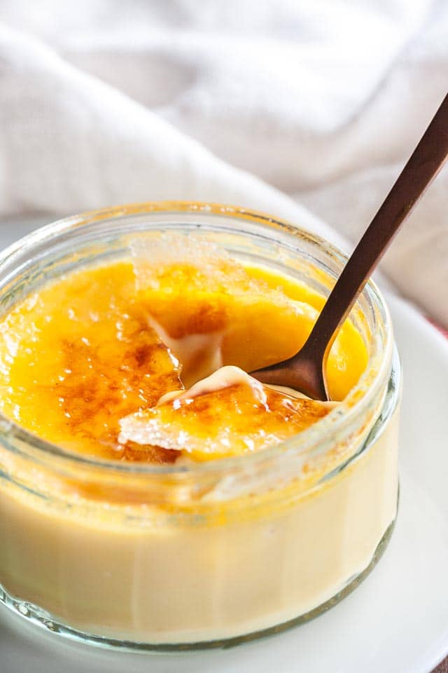 A glass of creme brulee with a bronze spoon cracking its top on a white plate in front of a white dishtowel.