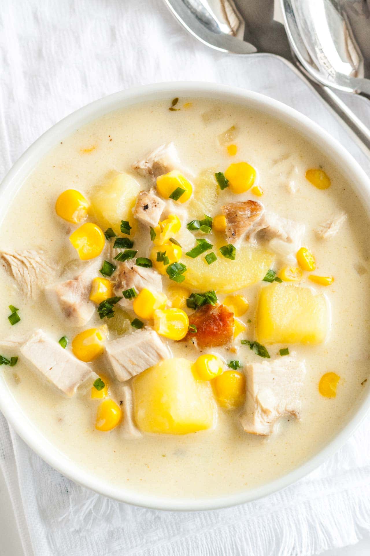 Top-down shot of a white bowl of creamy leftover turkey soup garnished with chives on a white dishtowel with two spoons next to it.