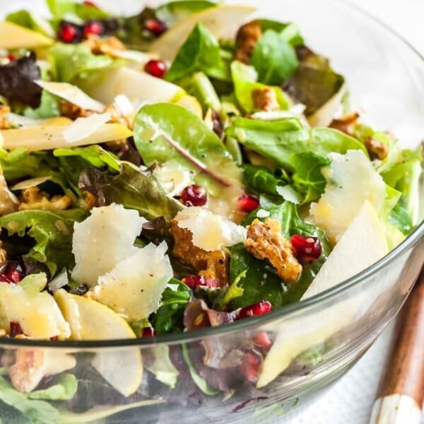 Close-up of a glass bowl with a pomegranate pear salad with walnuts on a white dish towel with wooden salad utensils.