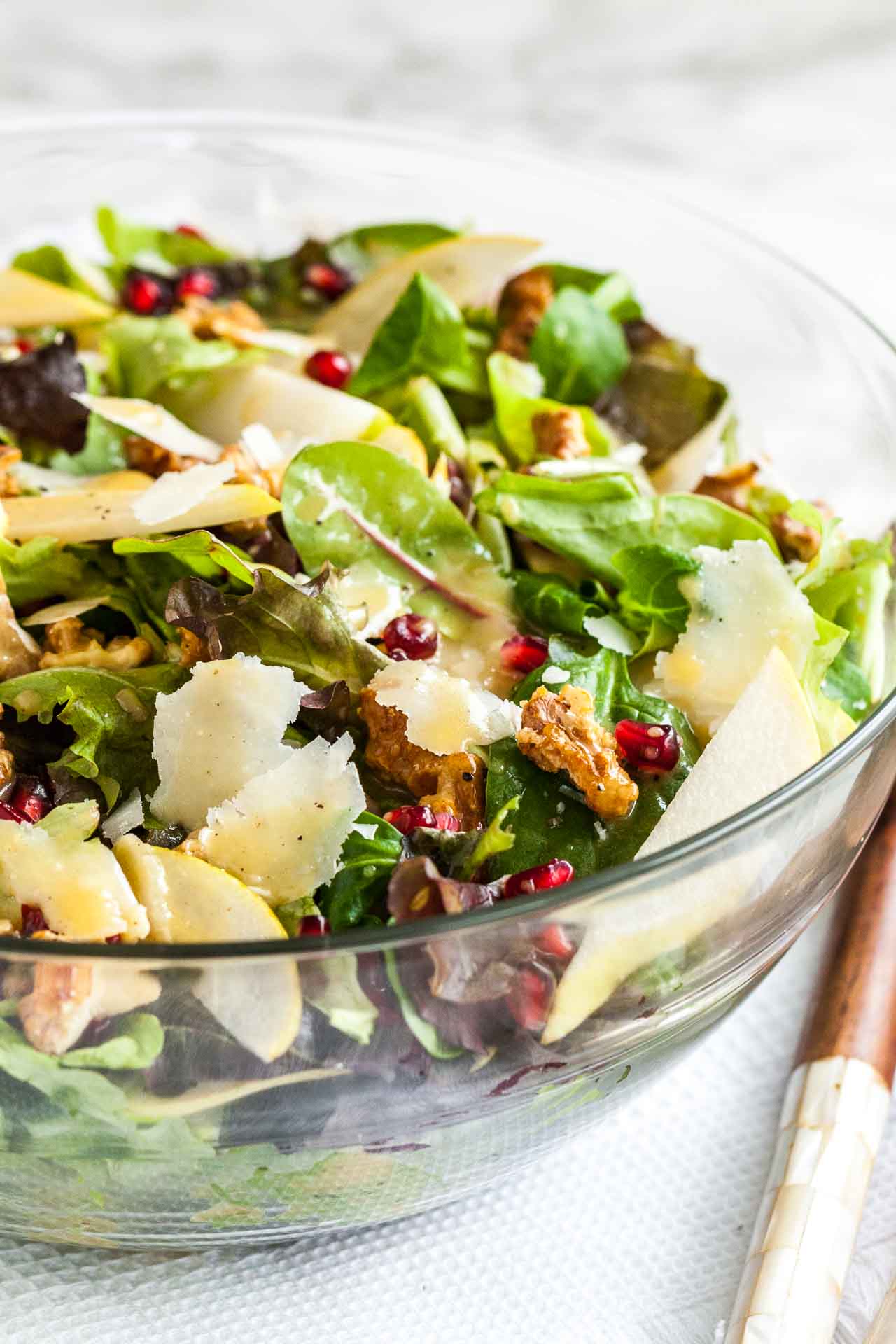 Close-up of a glass bowl with a pomegranate pear salad with walnuts on a white dish towel with wooden salad utensils.