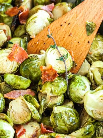 Close-up of roasted brussels sprouts with bacon and a sprig of thyme with a wooden cooking spatula in it.