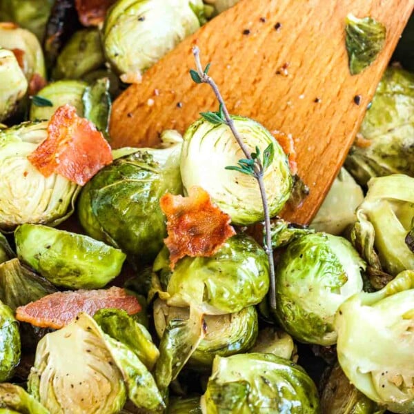 Close-up of roasted brussels sprouts with bacon and a sprig of thyme with a wooden cooking spatula in it.