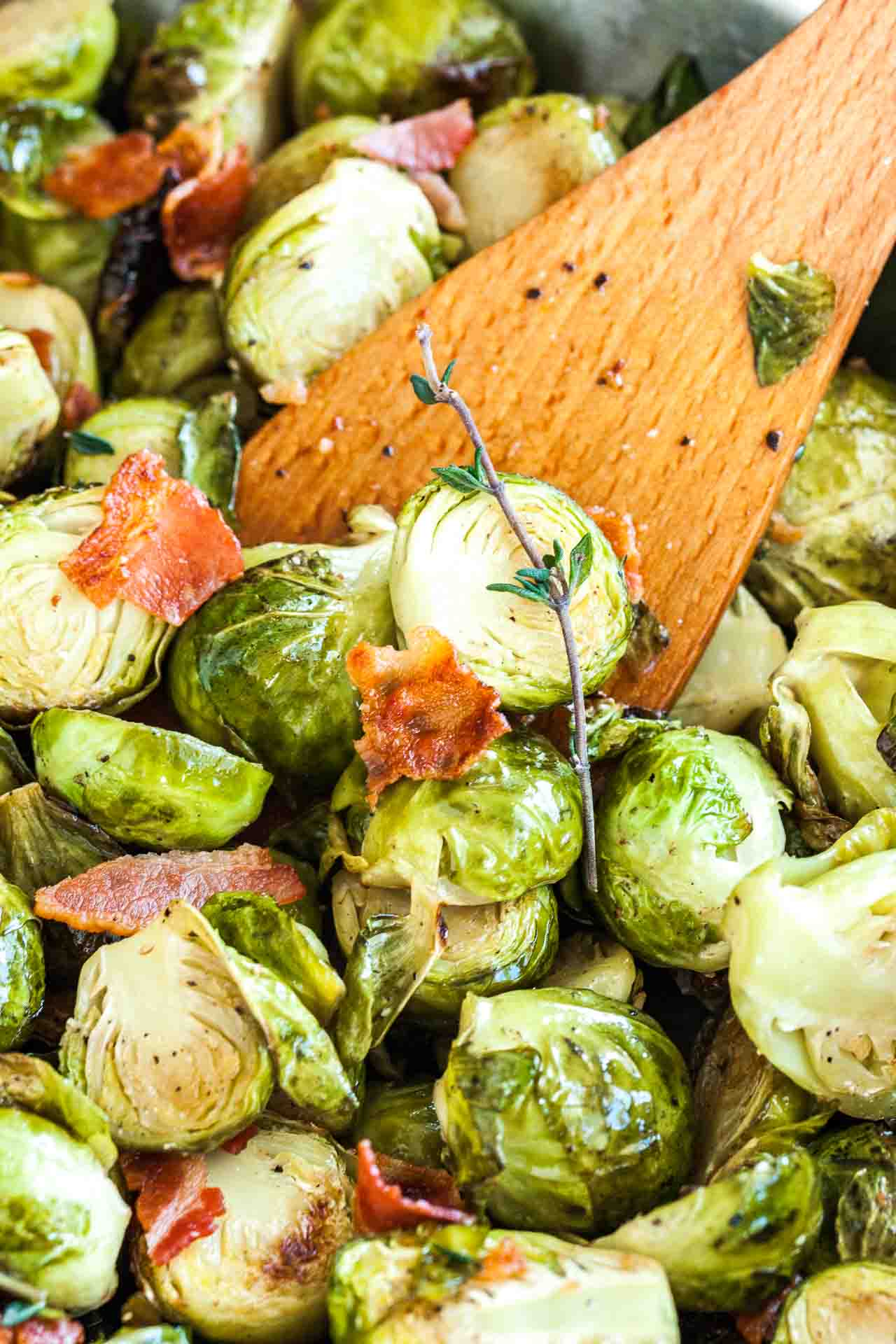 Close-up of roasted brussels sprouts with bacon and a sprig of thyme with a wooden cooking spatula in it.