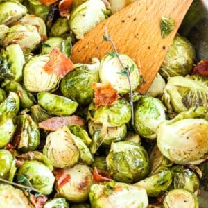 Roasted brussels sprouts with bacon and a sprig of thyme with a wooden cooking spatula in it in a stainless steel pan.