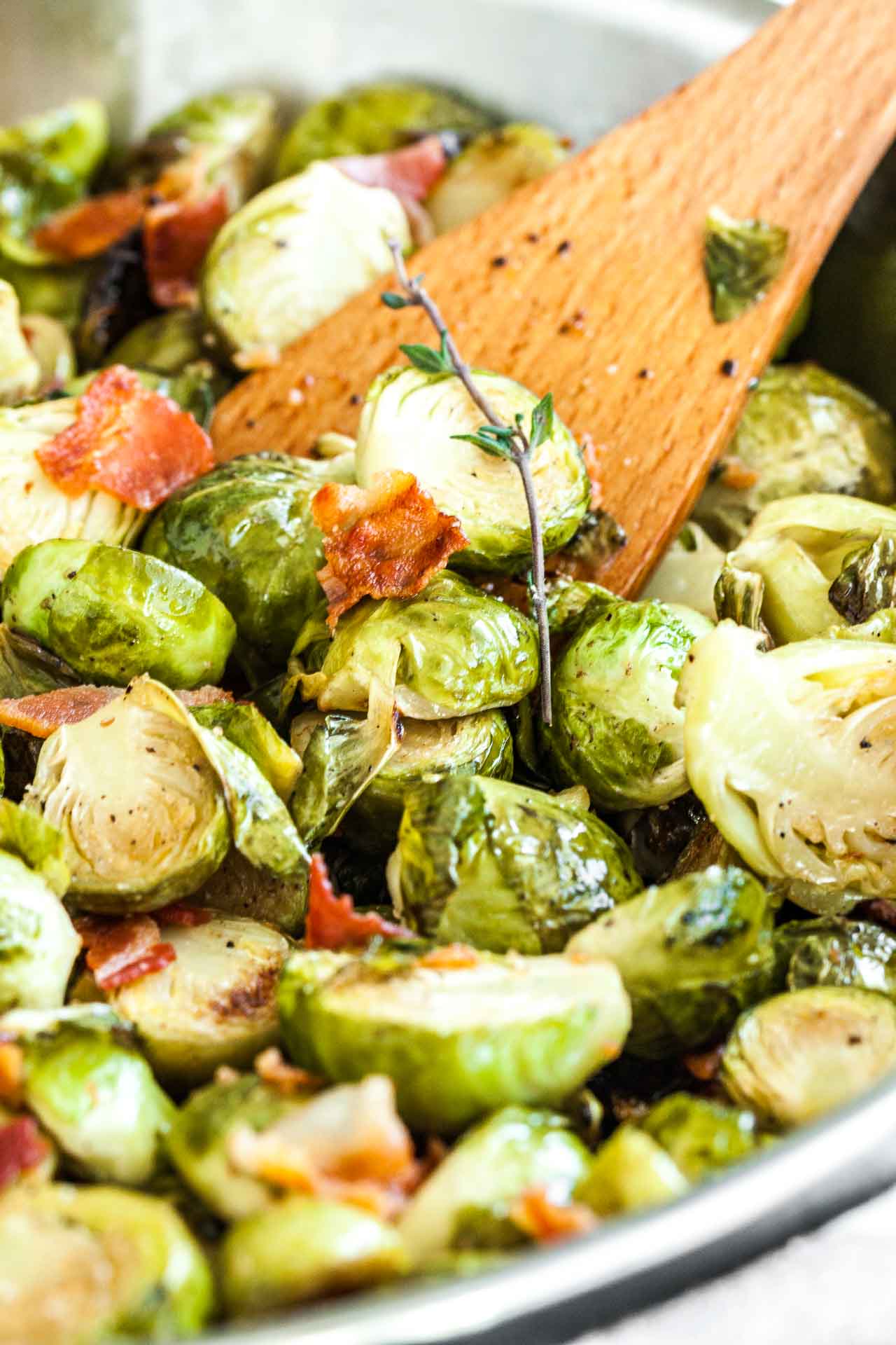 Close-up of roasted brussels sprouts with bacon and a sprig of thyme with a wooden cooking spatula in it.