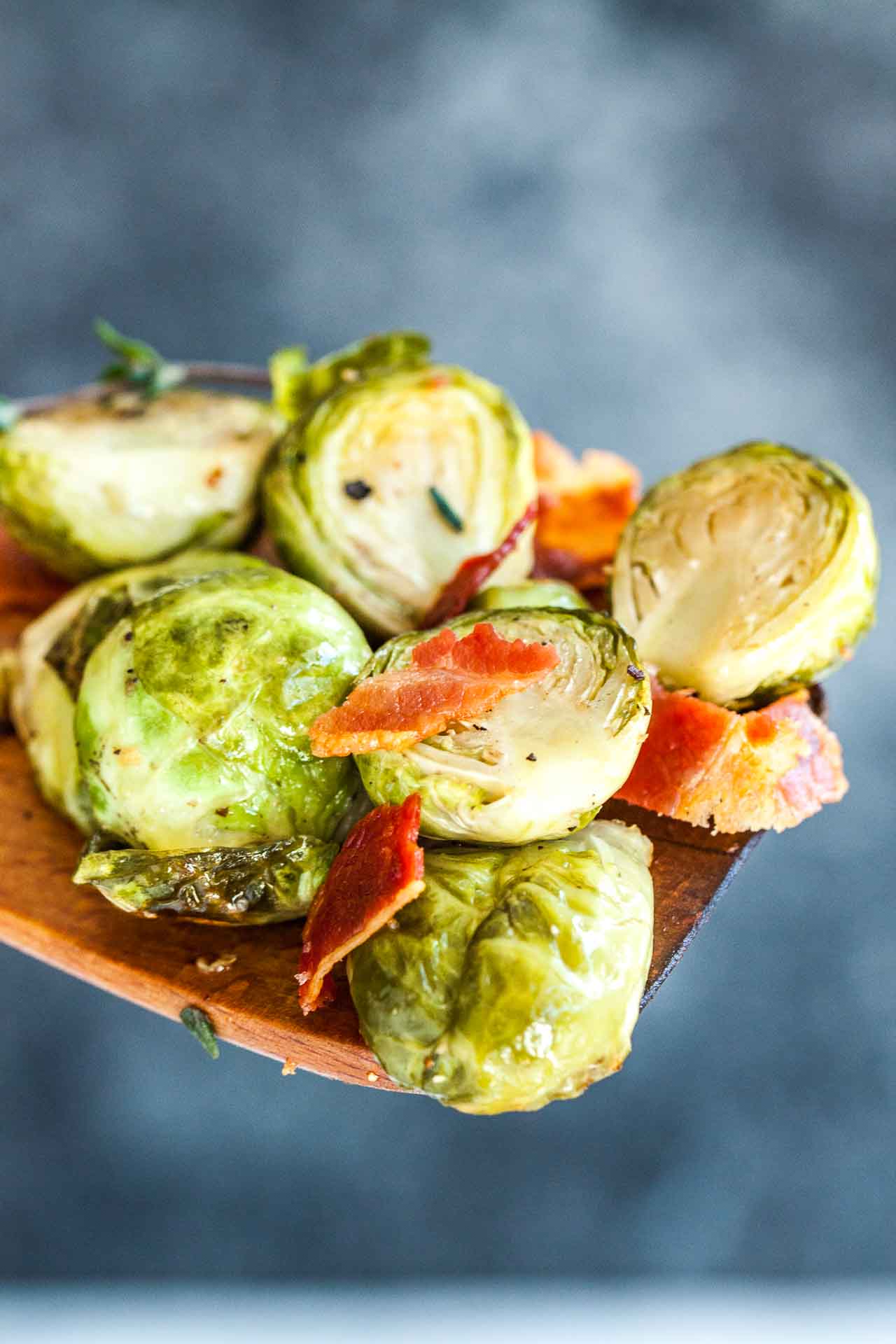 Roasted brussels sprouts with bacon on a wooden cooking spatula in front of a grey background.