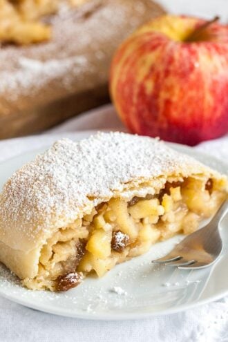 Close-up of a piece of apple strudel topped with powdered sugar on a white plate with a fork on a white dishcloth, with an apple in the background.